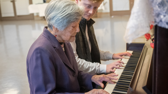 An older lady sits beside a younger man and plays the piano.