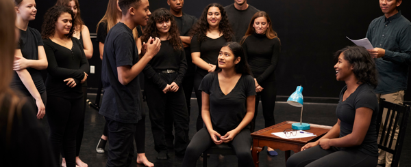 A group of ethnically diverse young people who are dressed in black, rehearse a scene from a play.
