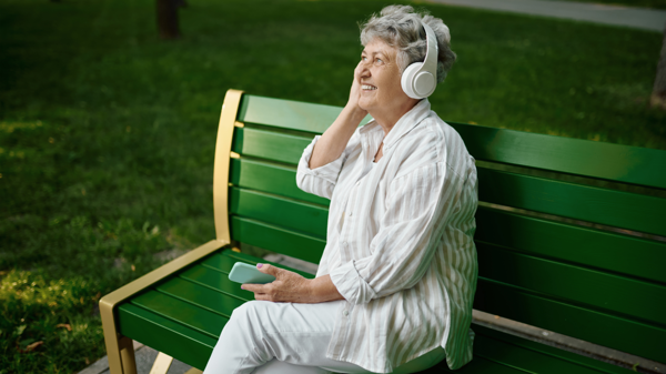 A woman sits on a park bench, she is listening to music in her headphones and smiling