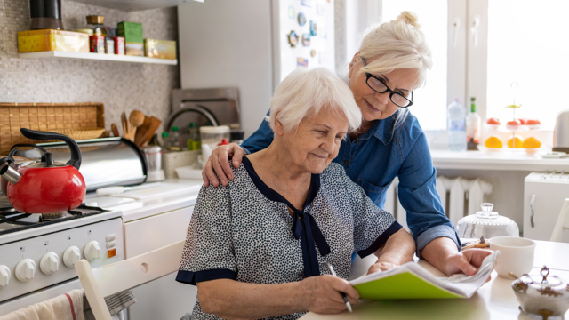 A daughter helps her older mother complete paperwork in the kitchen