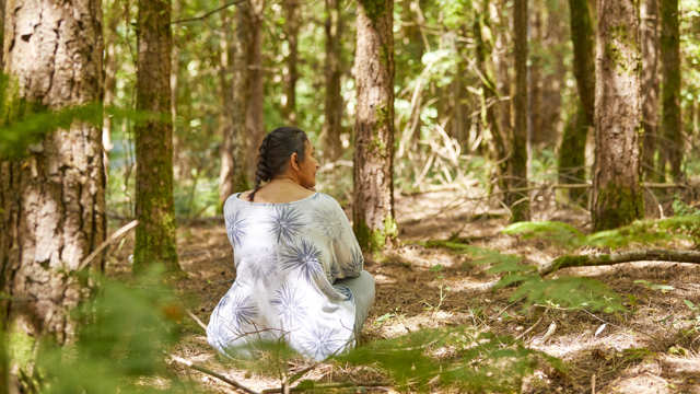 A woman sits peacefully, cross-legged on the ground among pine trees