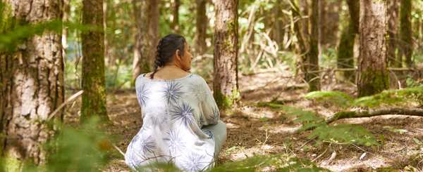 A woman sits peacefully, cross-legged on the ground among pine trees