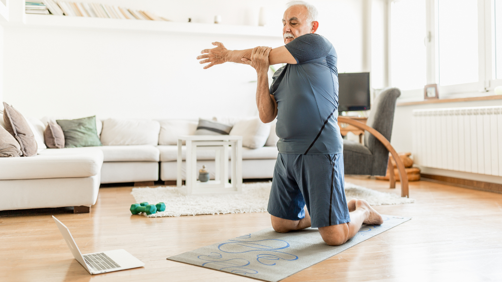An older man look at a laptop and stretches his arm, while taking part in an online exercise class from his living room