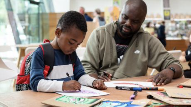 A Black father and young boy draw together in an art gallery