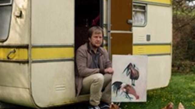 A man sitting on the step of a caravan holds up a painting of cockerels he has made as part of the Art by Post initiative 