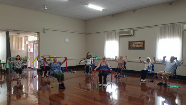 A group of older adults sit in chairs holding up exercise equipment