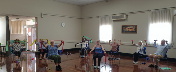 A group of older adults sit in chairs holding up exercise equipment