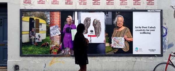 A woman looks at a billboard of the Art by Post initiative. The billboard shows participants and their art works alongside a caption that reads: Art by Post: Unlock creativity for your wellbeing