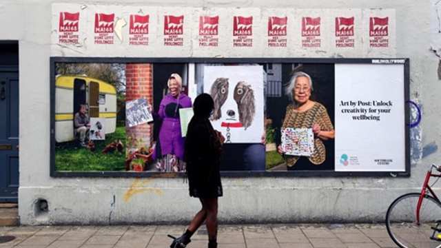 A woman looks at a billboard of the Art by Post initiative. The billboard shows participants and their art works alongside a caption that reads: Art by Post: Unlock creativity for your wellbeing