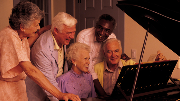 Three older adults stand around an older couple playing a piano. 