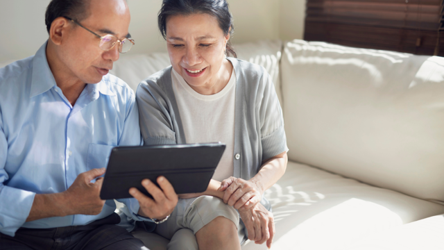 An older Asian man and woman sit on a sofa and read a tablet