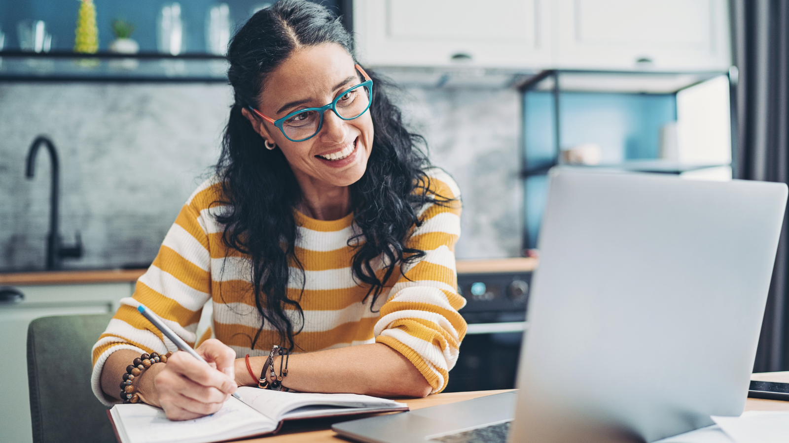 A young woman looks and smiles at a laptop screen while writing notes in a notebook