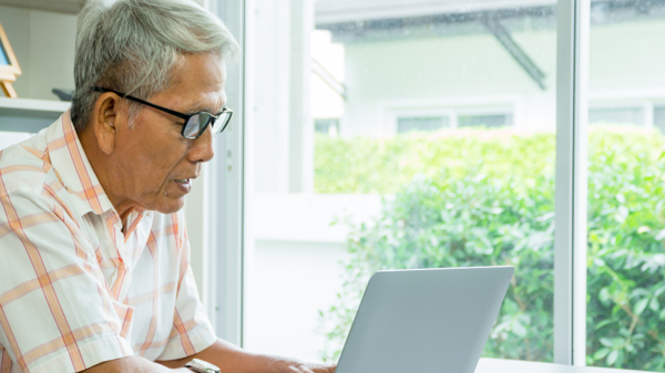 An older man sits at a table and looks at a laptop