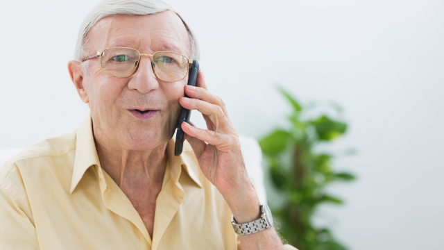An older white man with glasses holds a mobile phone to his ear and speaks