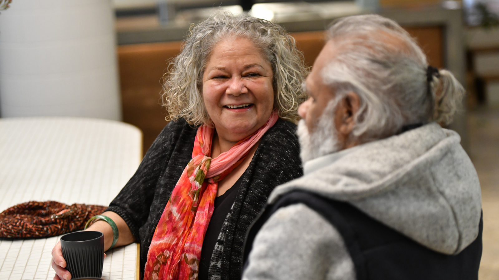 A white women smiles at an Asian man while holding her coffee cup and sitting at a table.