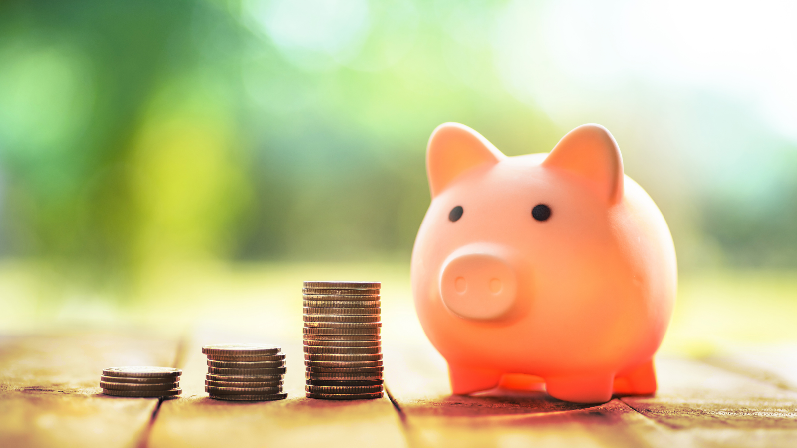 Pink pig in the shape of a piggy bank with three stacks of coins next to it, sitting on a wooden table. 