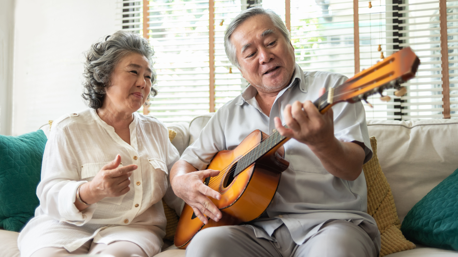 A couple are playing the guitar together on a sofa, they are singing and smiling