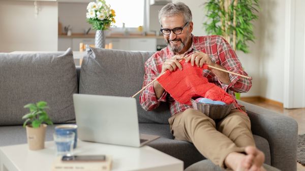 A man sits on a sofa and knits red wool while smiling at a laptop screen