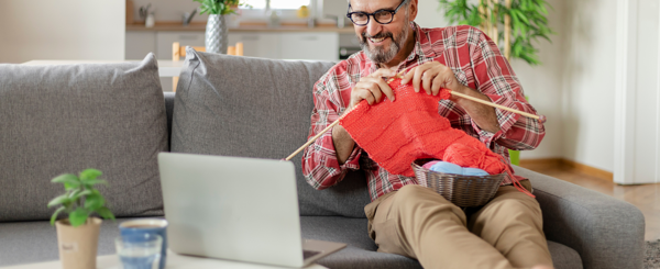 A man sits on a sofa and knits red wool while smiling at a laptop screen