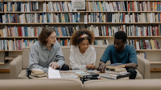 Three young adults sit on a sofa in a library and read a book