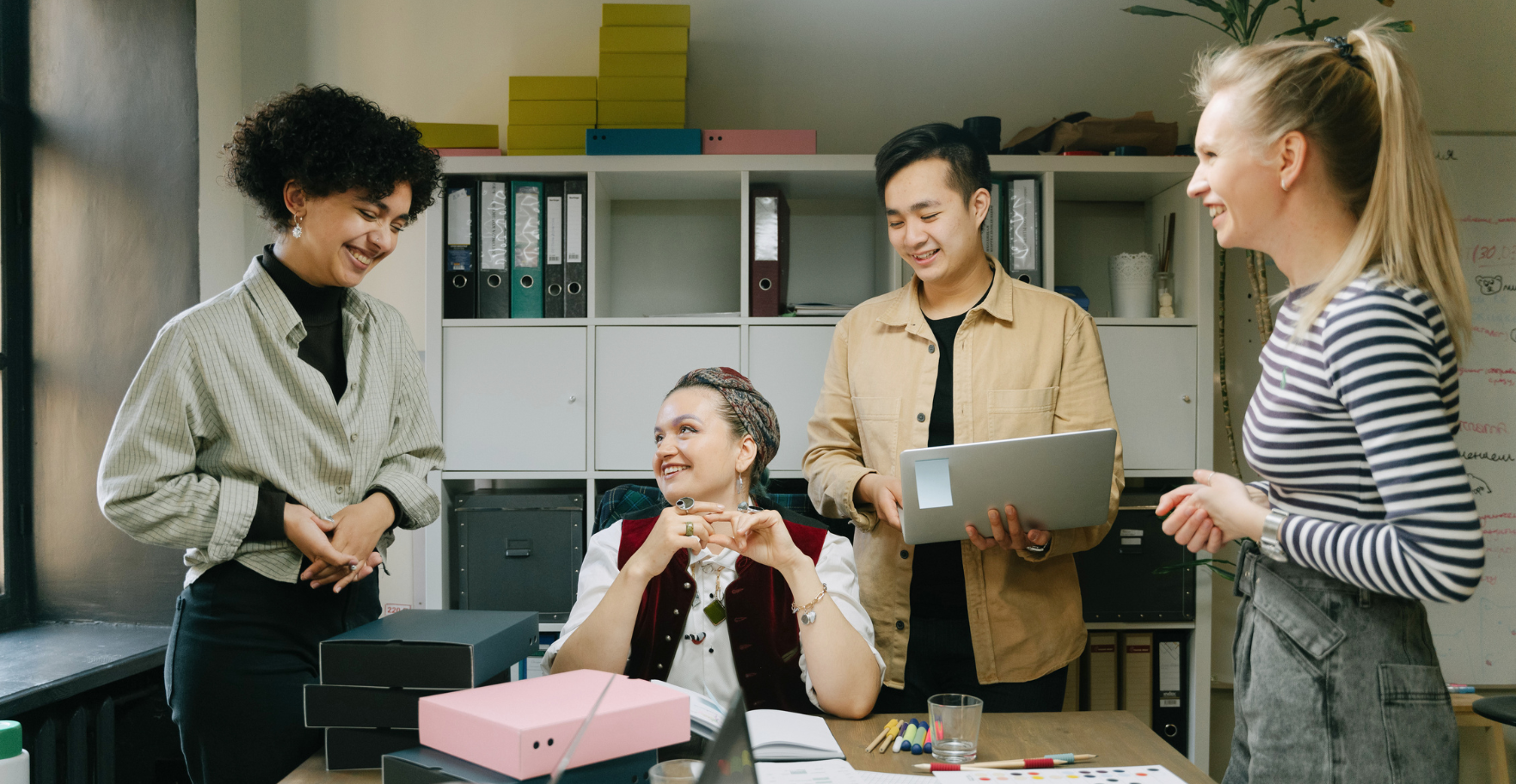 Office workers stood and sat around a desk with files and laptops, smiling and talking