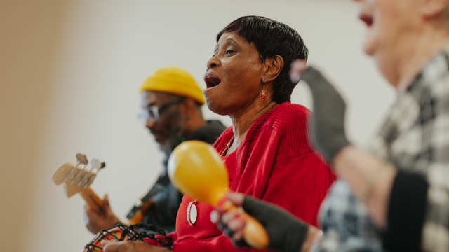 A group of adults sit in a line and play the guitar, drums and maracas. 