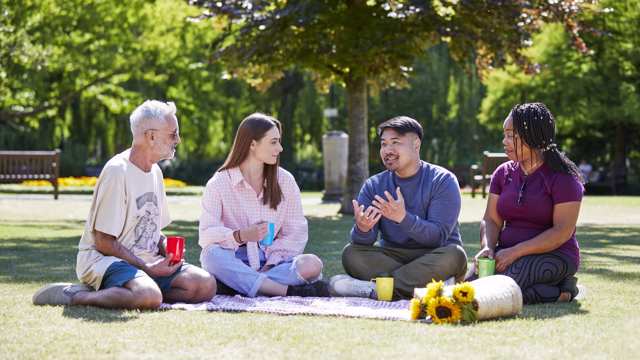 Four people of varying ages and ethnicities sit around a picnic blanket in a park, drinking from mugs and talking
