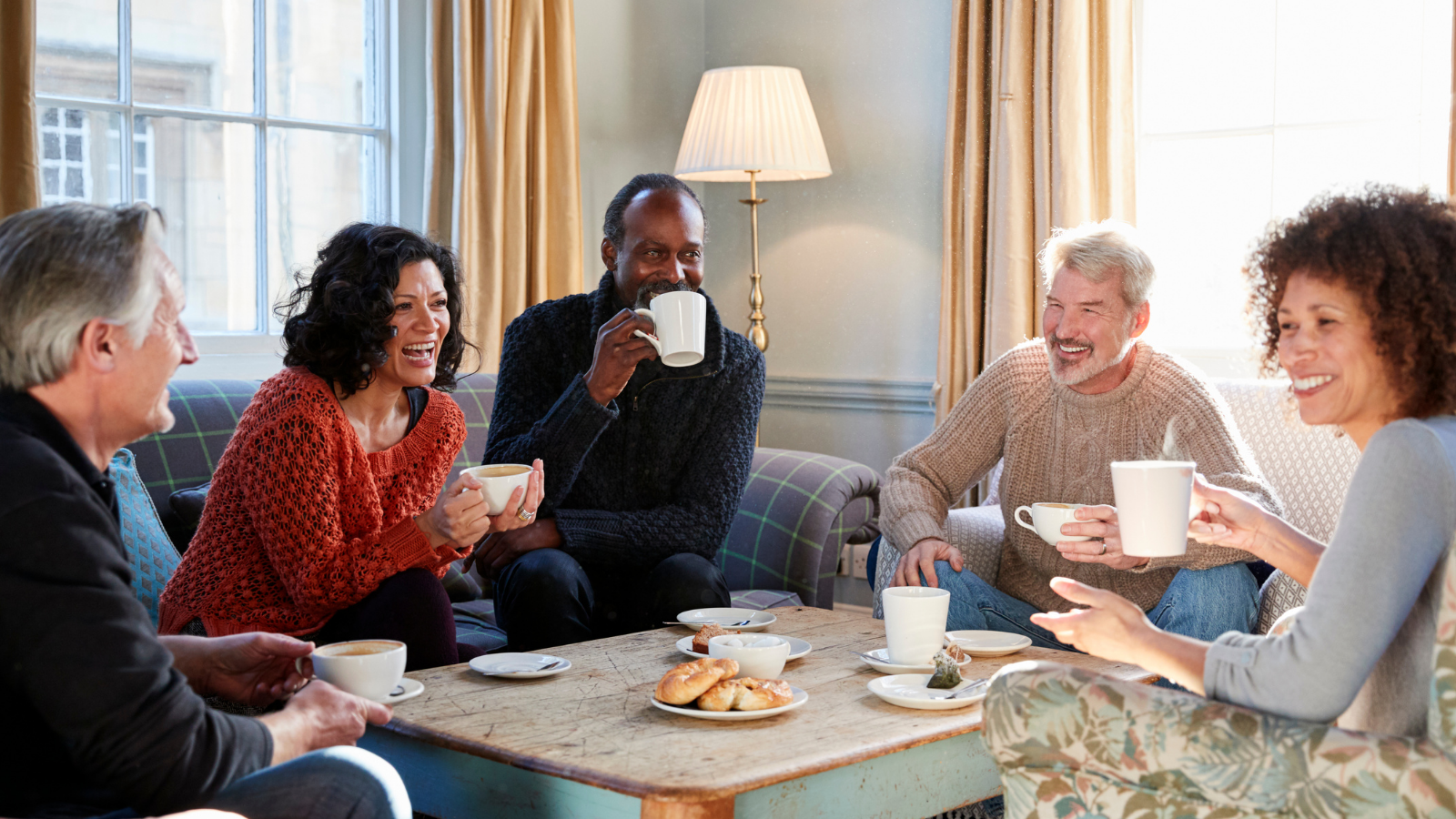 A group of adults of varying ethnicities sit around a table and laugh while drinking tea and eating pastries