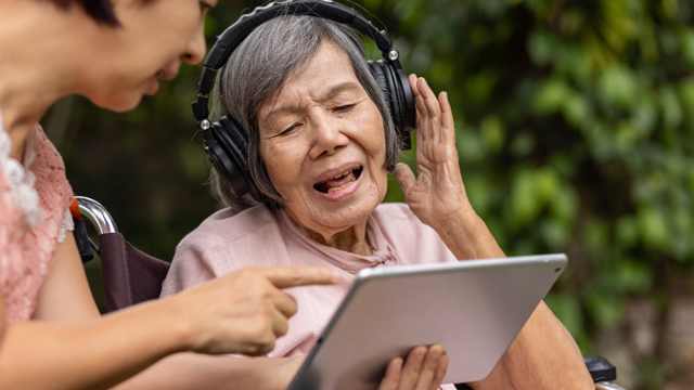 An older Asian women in a wheelchair wears headphones and listens to music on a tablet held up by another woman