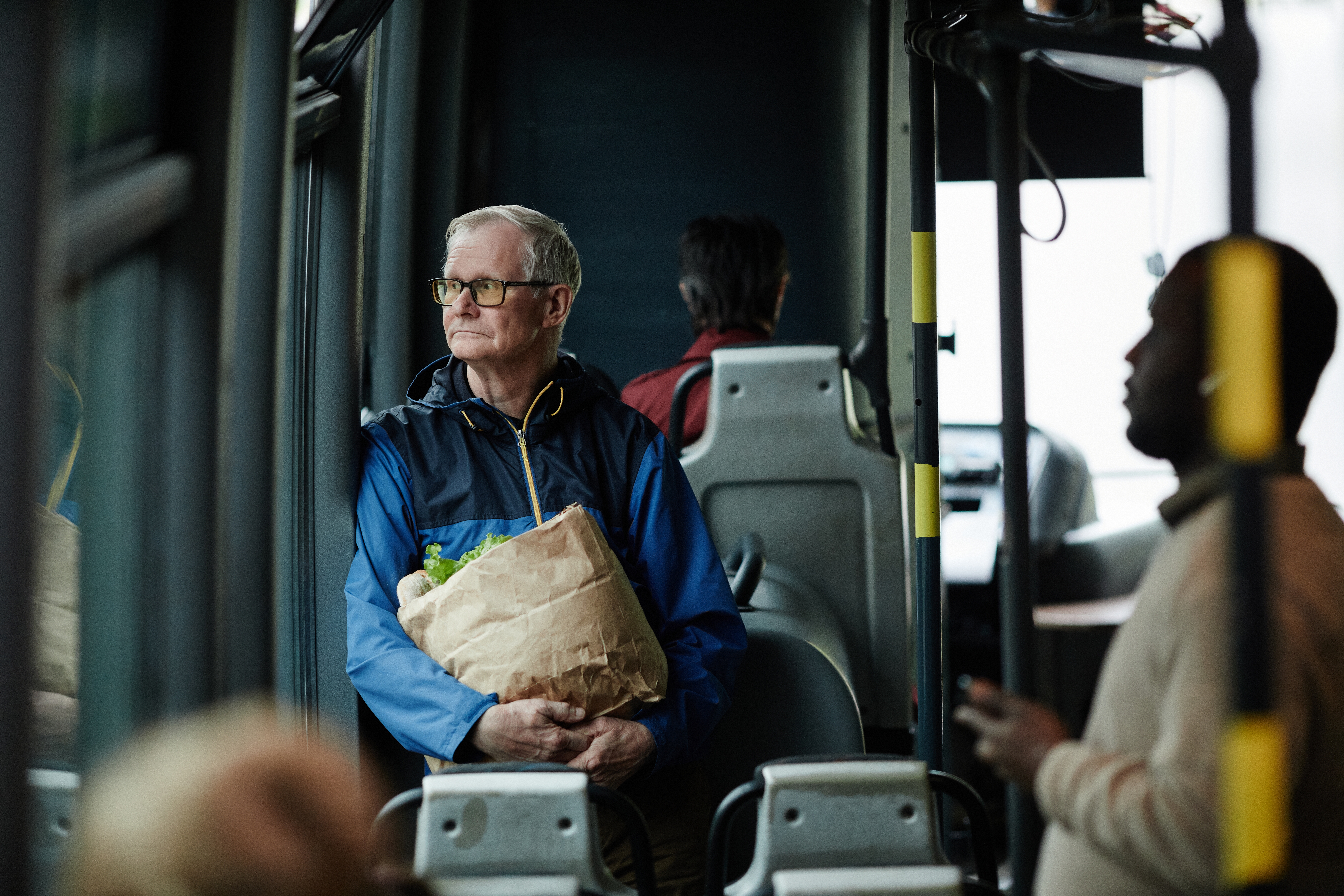 An older man sits alone on a bus staring sadly out of the window holding a bag of shopping 