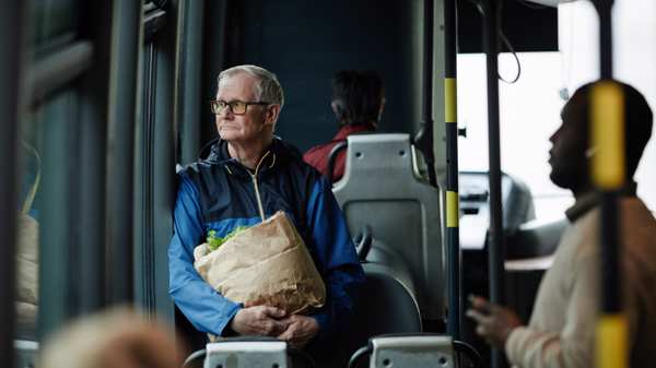 An older man sits alone on a bus staring sadly out of the window holding a bag of shopping 
