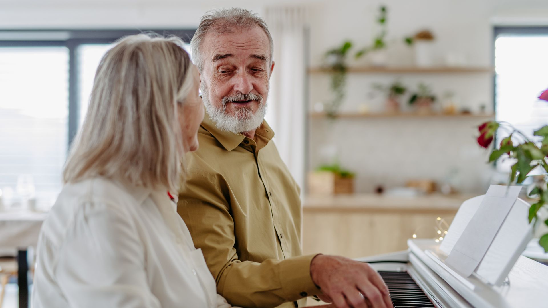 An older lady and gentleman sitting at a piano together, he is showing her the notes and they are smiling