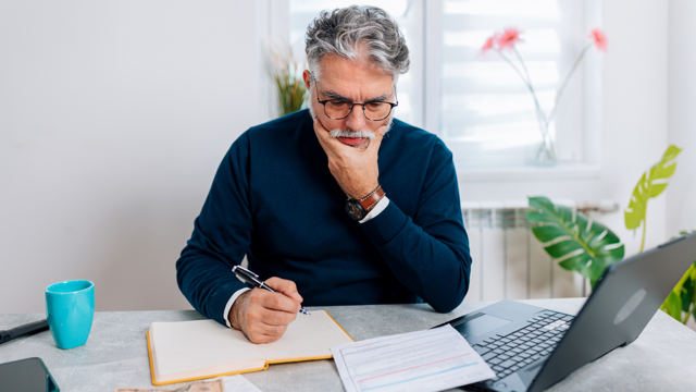 A man sits at a table with bills and a laptop and writes into a notebook