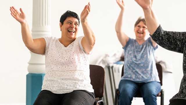 Two middle aged women smile as they sit in chairs with their arms in the air