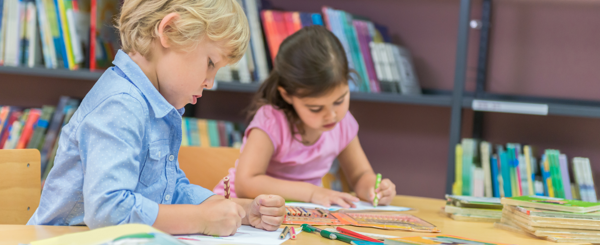 Two children sit at a table at a library and colour drawings 