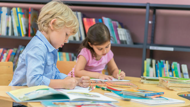 Two children sit at a table at a library and colour drawings