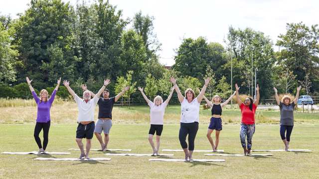 A group of people with their arms outstretched take part in an outdoor exercise class in a park