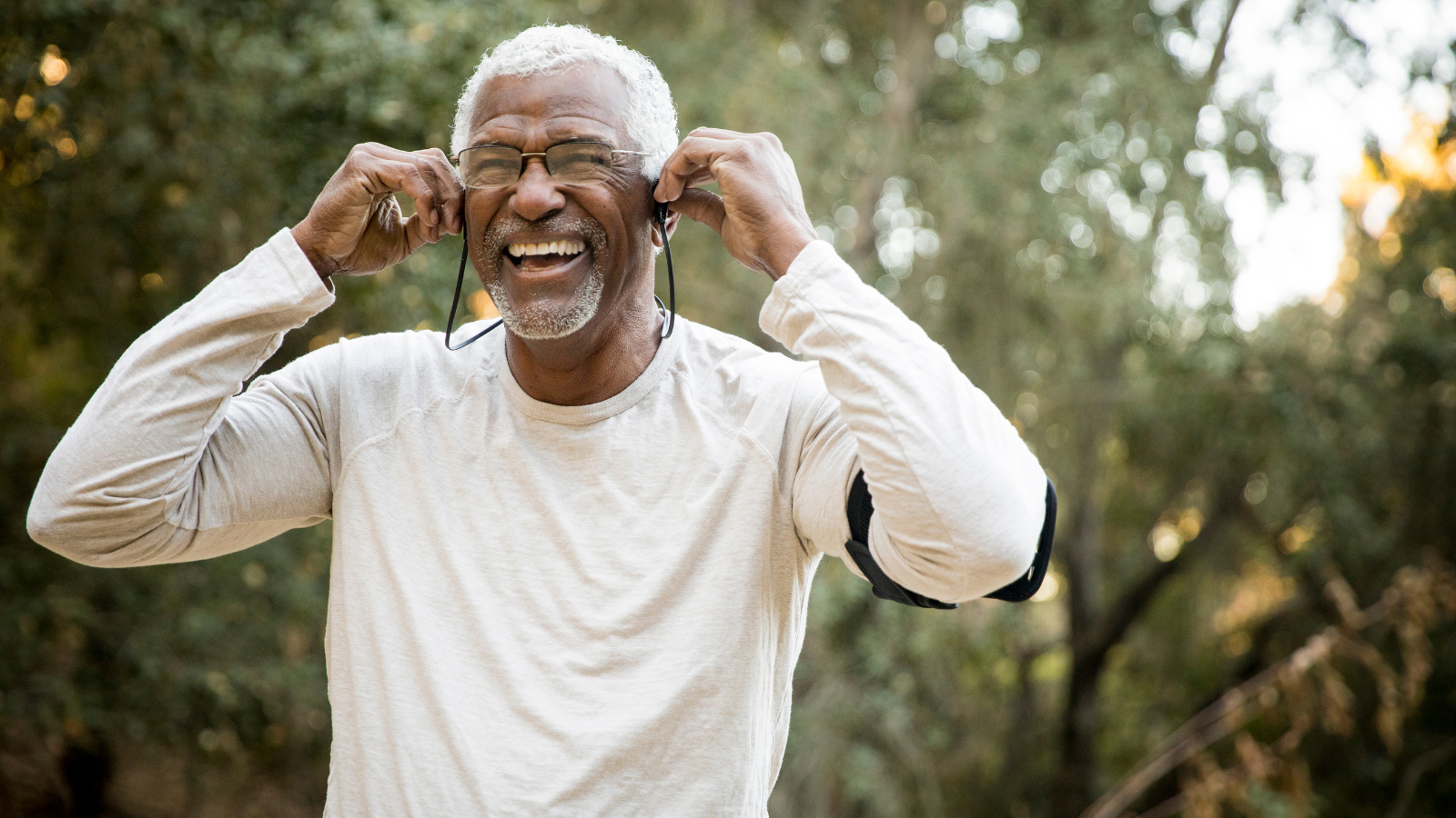 An older man stands in a park and smiles while listening to music on and adjusting his headphones