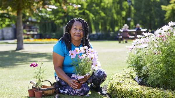 A woman sat on the grass in a park holding a plant she is about to put in the ground 