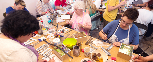 A large groups of adults sit around a table with colourful art supplies and paint