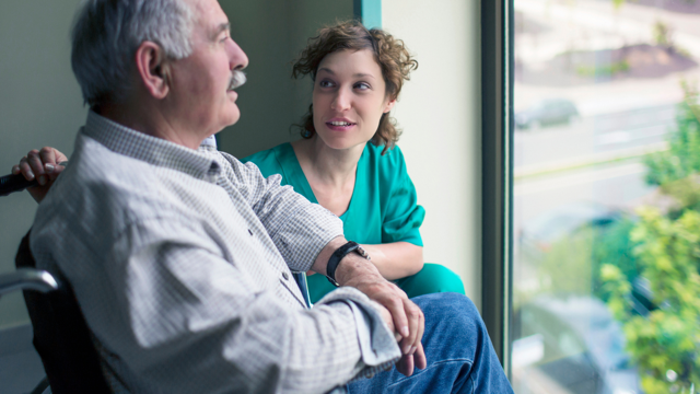A women faces and talks to an older man who is sitting in a wheelchair facing a window