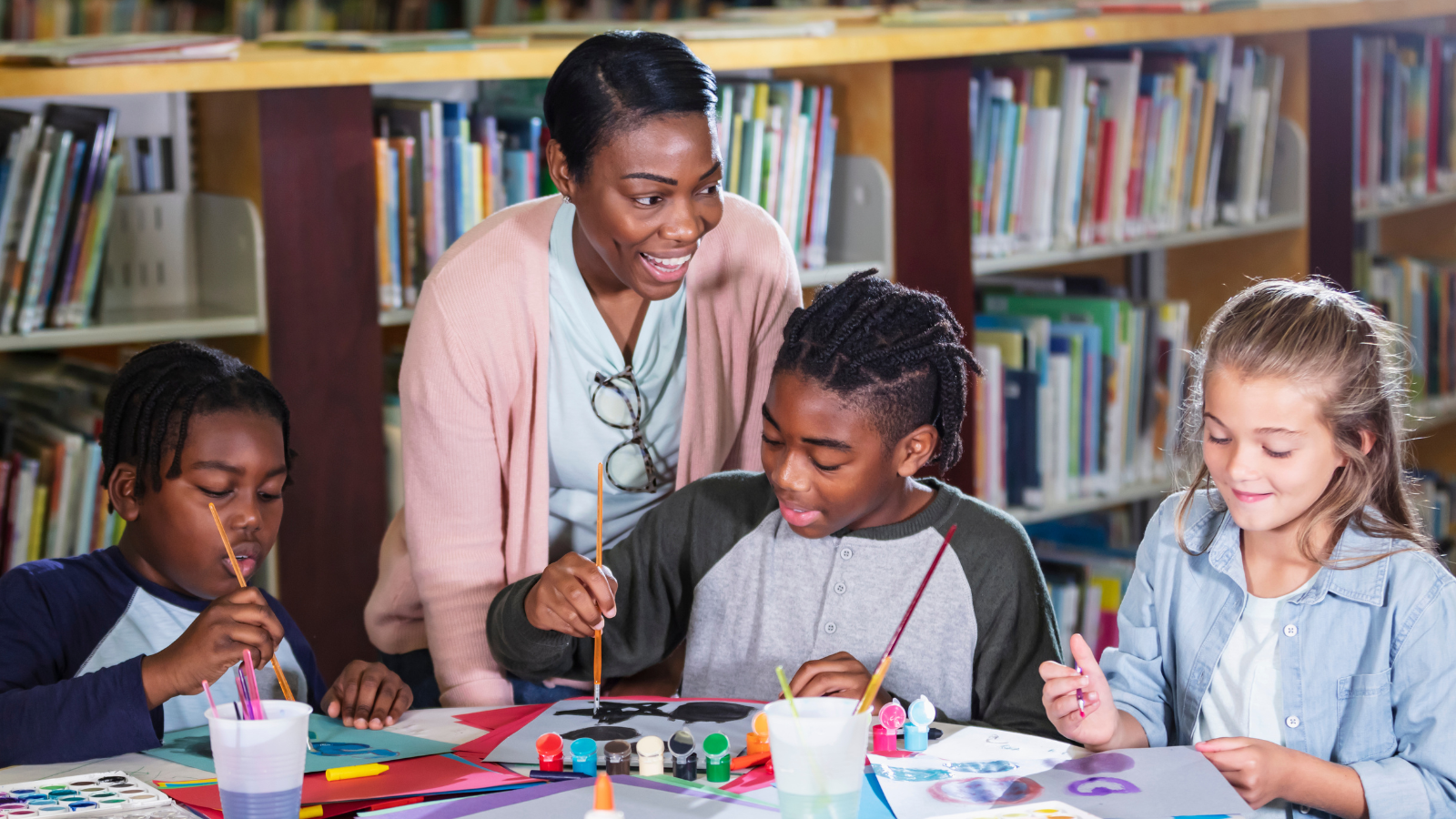 A group of children sit at a table in library and paint. A librarian watches over them.