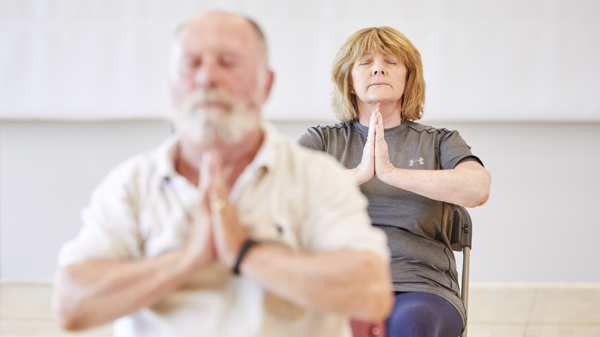 Two older people participate in chair yoga, they sit with their eyes closed and hands pressed together at their chest