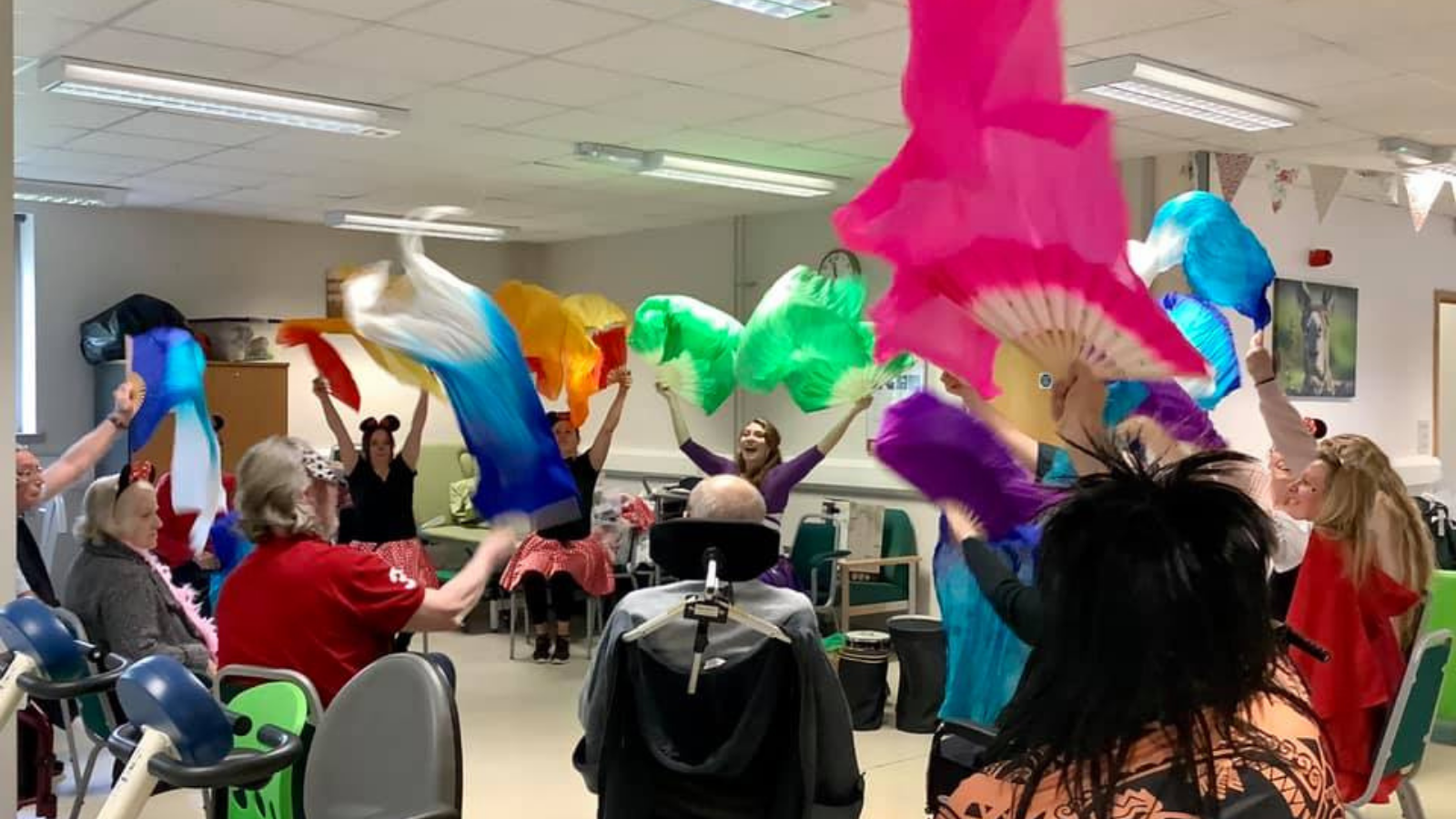 A group of variously abled older adults sit in a circle waving colourful silks above their heads