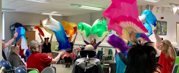 A group of variously abled older adults sit in a circle waving colourful silks above their heads