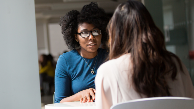 Two women talking. A Black woman is facing a women with 	long brown hair across a table