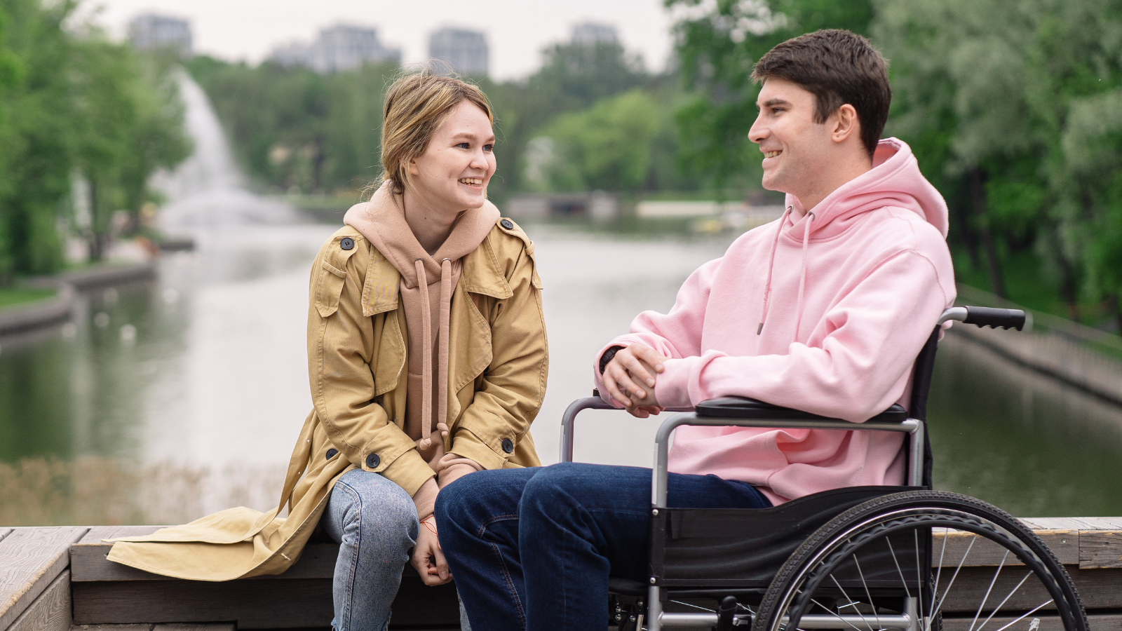 A young man in a wheelchair and a young woman chat while sitting on a bench on a riverfront