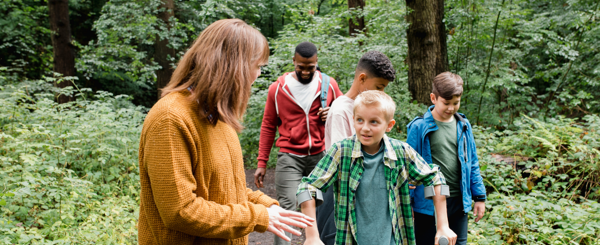 A group of children of varying ethnicities walk through a forest path with two teachers