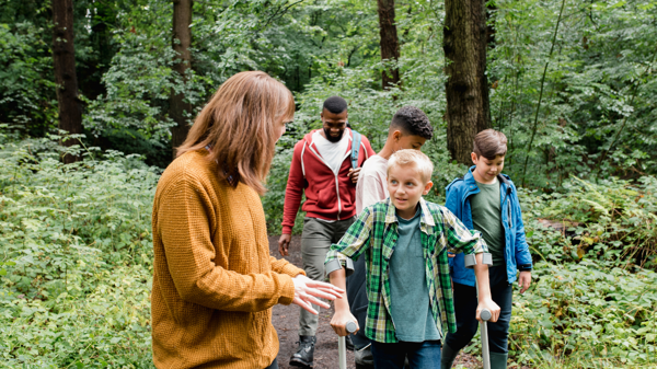 A group of children of varying ethnicities walk through a forest path with two teachers