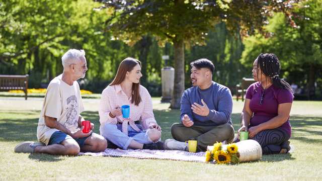 A group of four friends sit on a blanket in the park while chatting together and drinking tea 